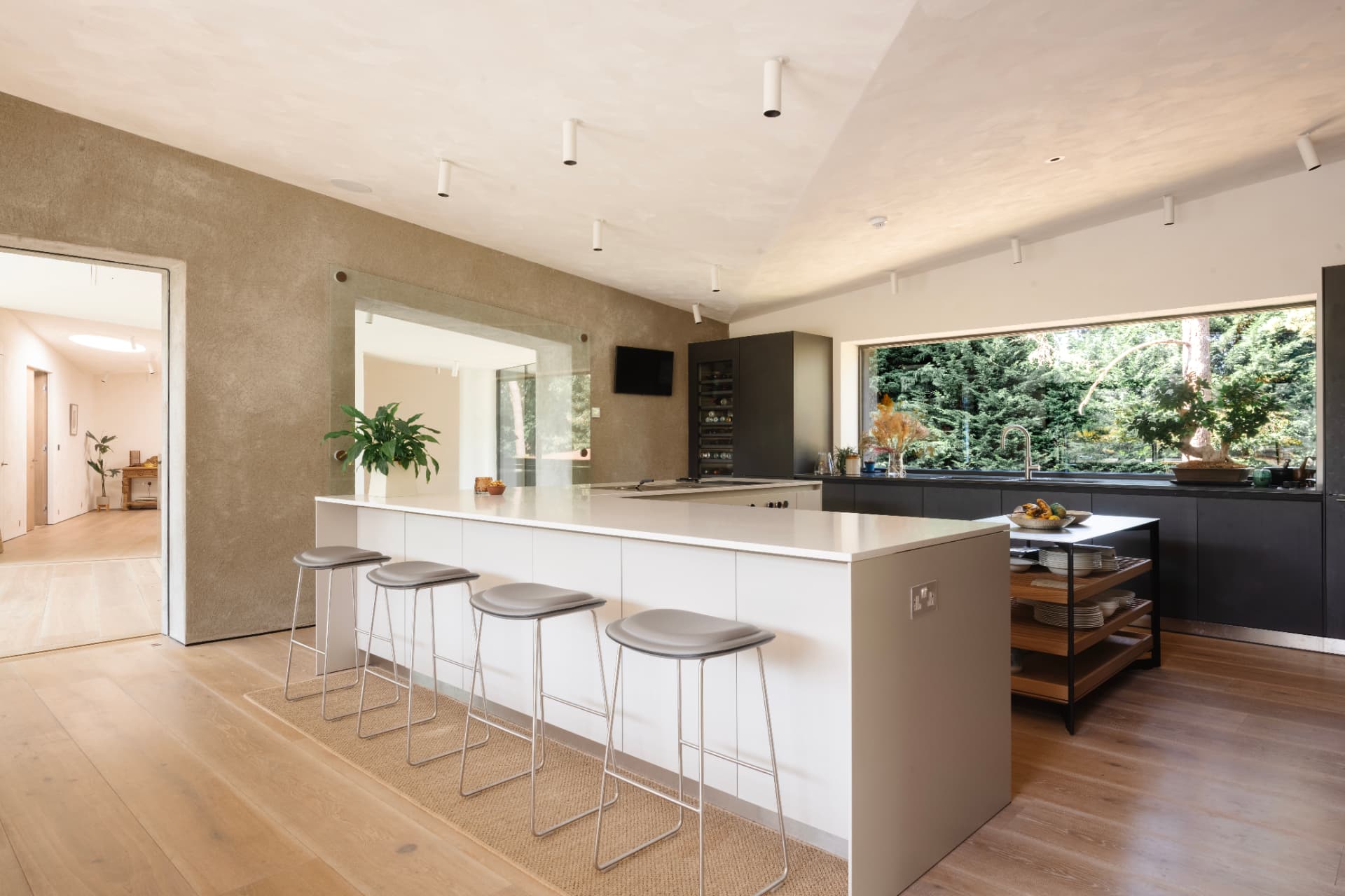 An open-plan kitchen with a white island and four stools, dark units along the rear wall and a wide window framing woodland
