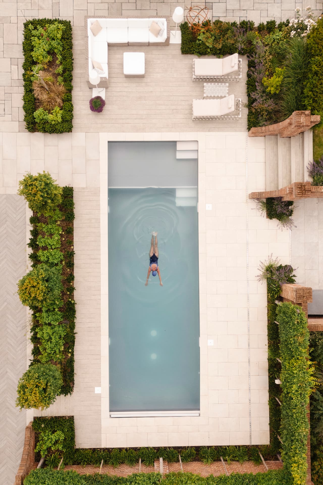 An overhead view of a swimmer crossing a long stone-edged pool set within clipped hedging, paving and terraced beds