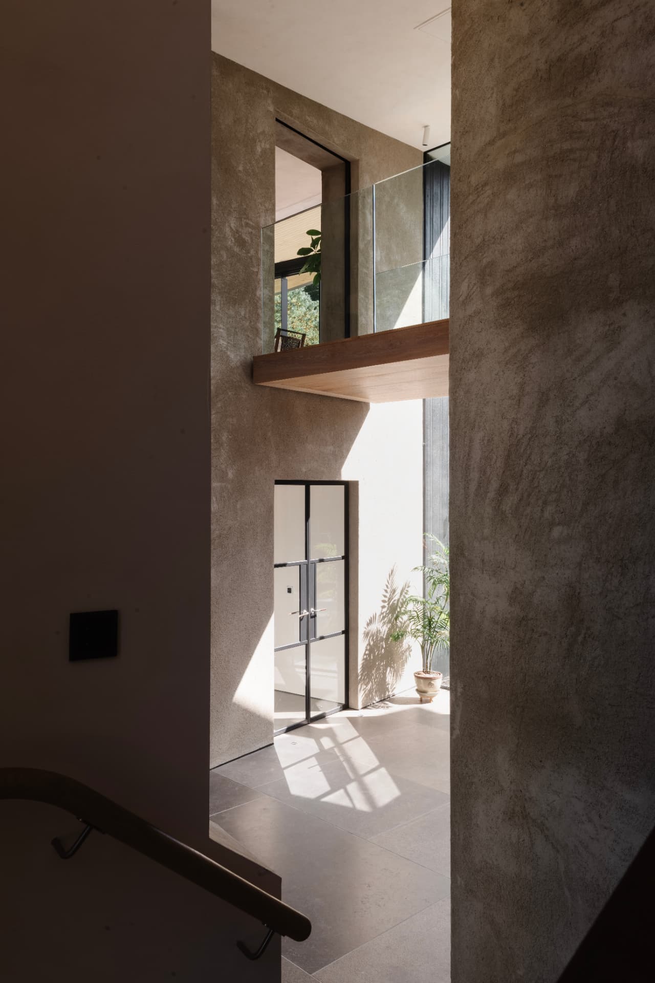 A double-height hall in polished plaster, with a timber gallery walkway above and sunlight falling across stone floor towards a glazed entrance door
