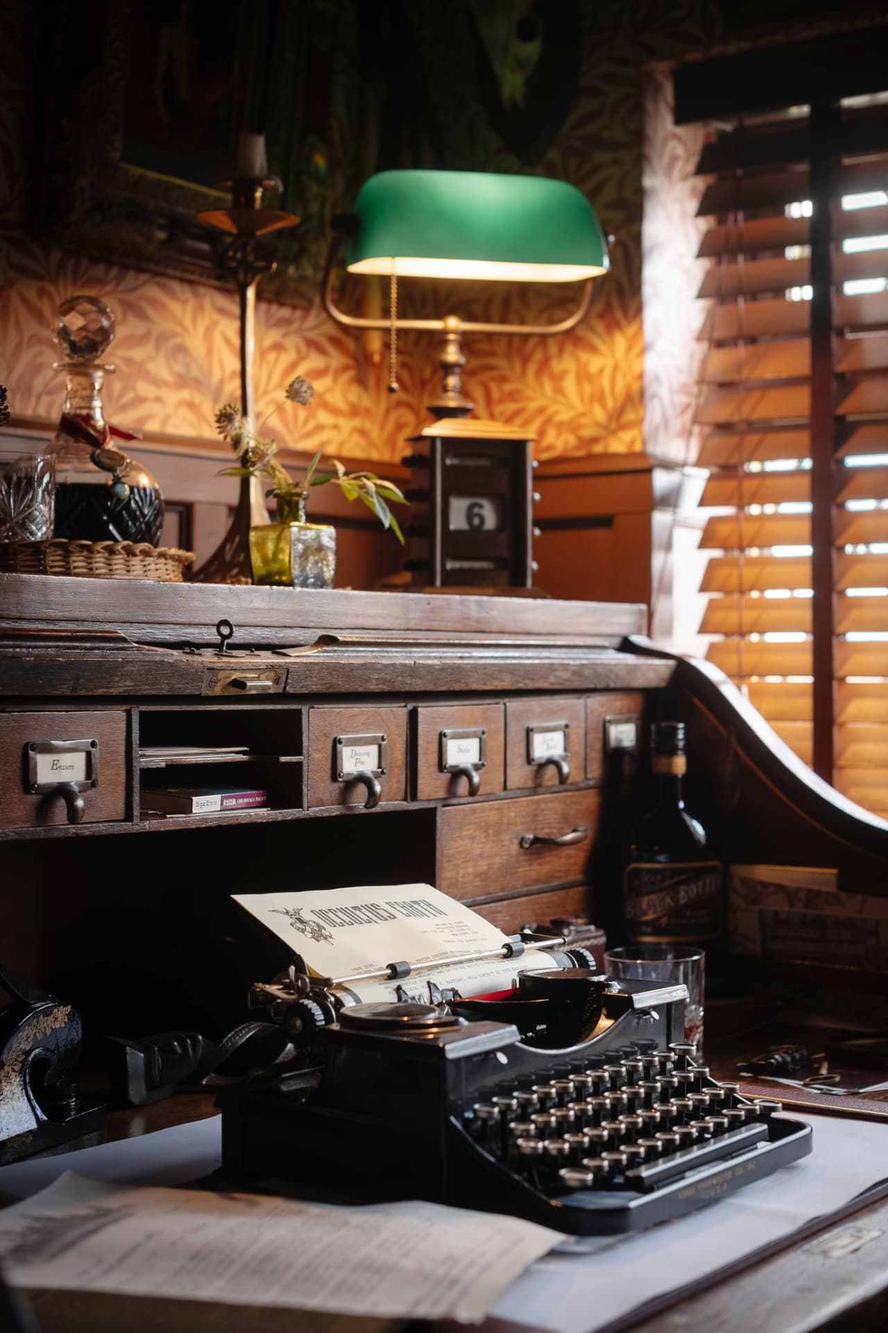 A vintage typewriter on an antique oak roll-top desk beneath a green banker's lamp, with patterned wallpaper and timber shutters behind