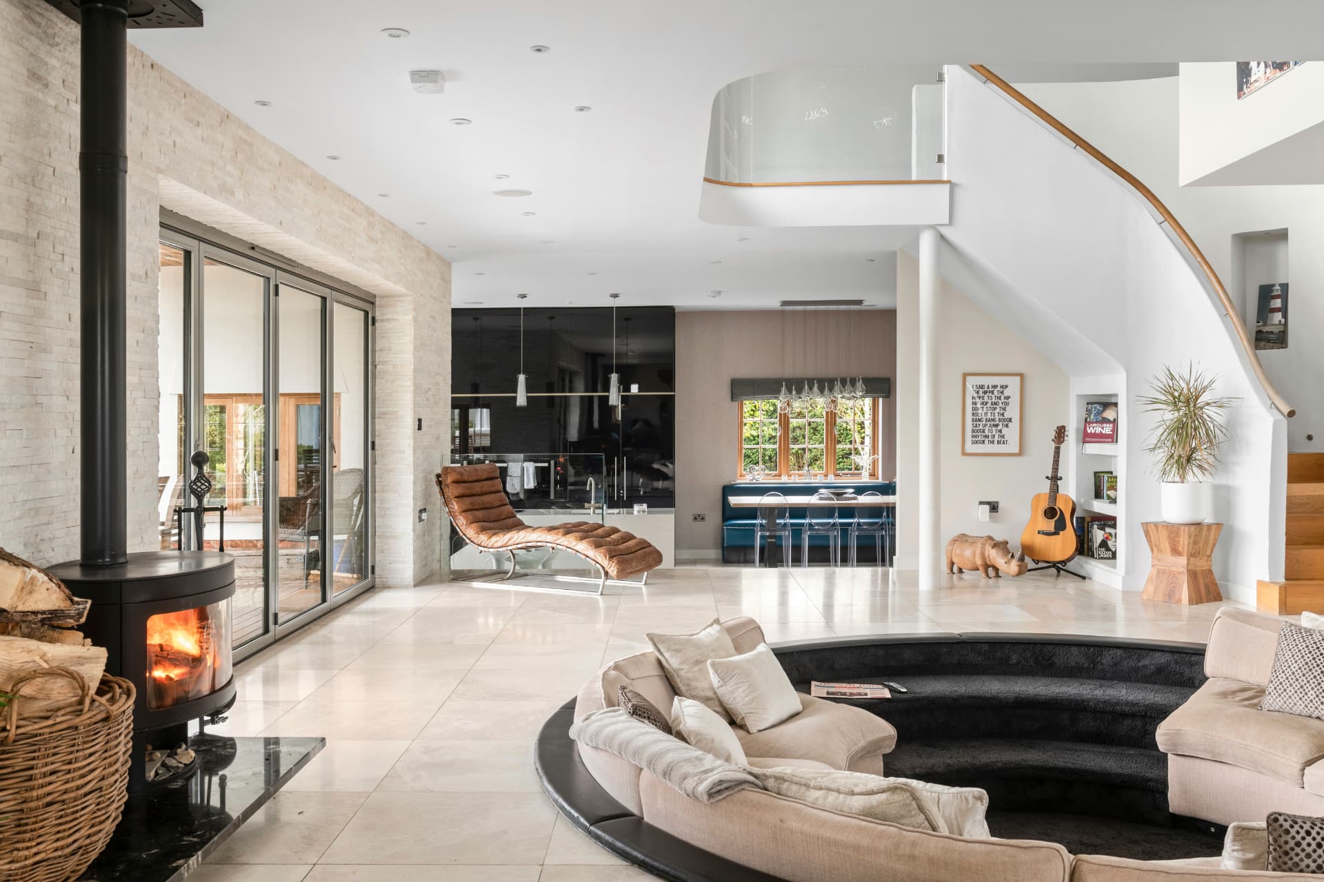 An open-plan living space with a wood-burning stove against split-stone walling, a sunken seating well, curved staircase and bifold doors to a deck