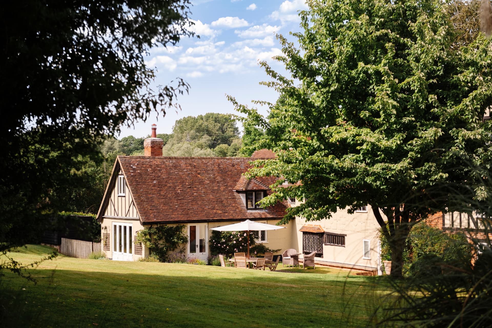 A timber-framed cream cottage with a clay tile roof and brick chimney, framed by trees across a sunlit lawn with garden furniture and a parasol