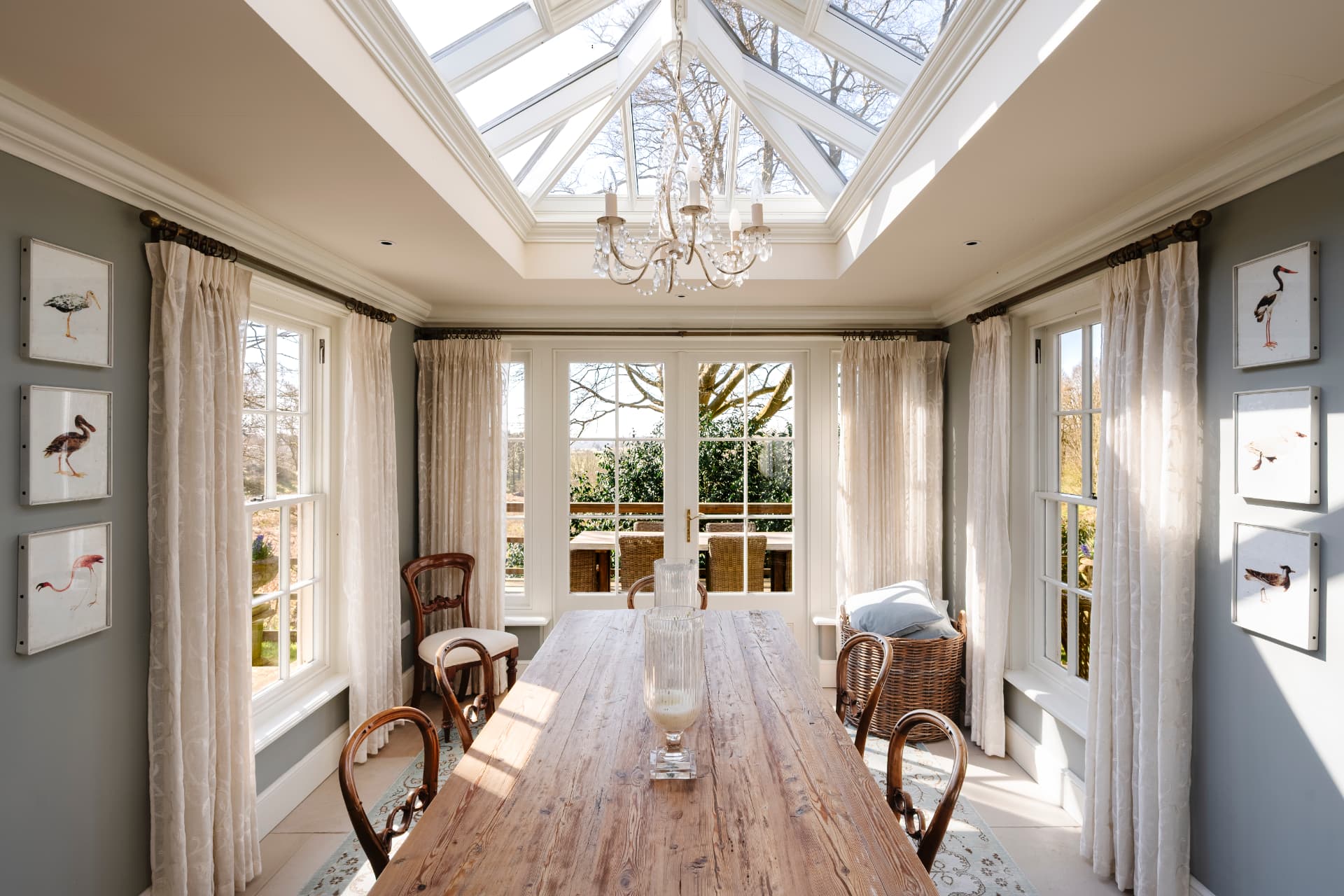 An orangery dining room under a glazed lantern roof and crystal chandelier, with a scrubbed pine table, bentwood chairs and framed bird studies on grey-green walls