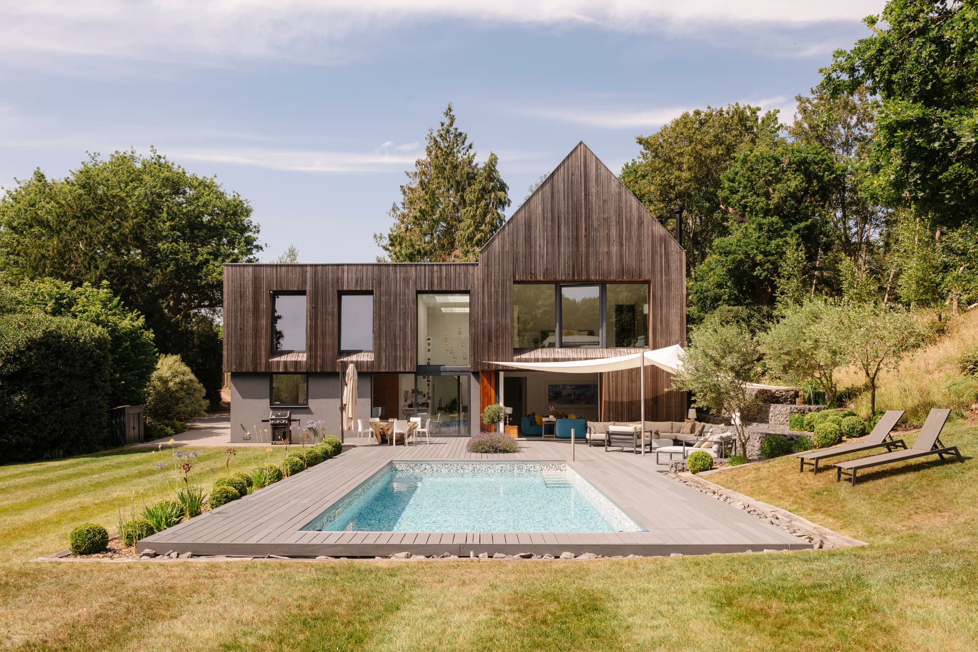 A dark timber-clad house with a steeply pitched gable, seen across a lawn and a rectangular swimming pool with decked surround and loungers