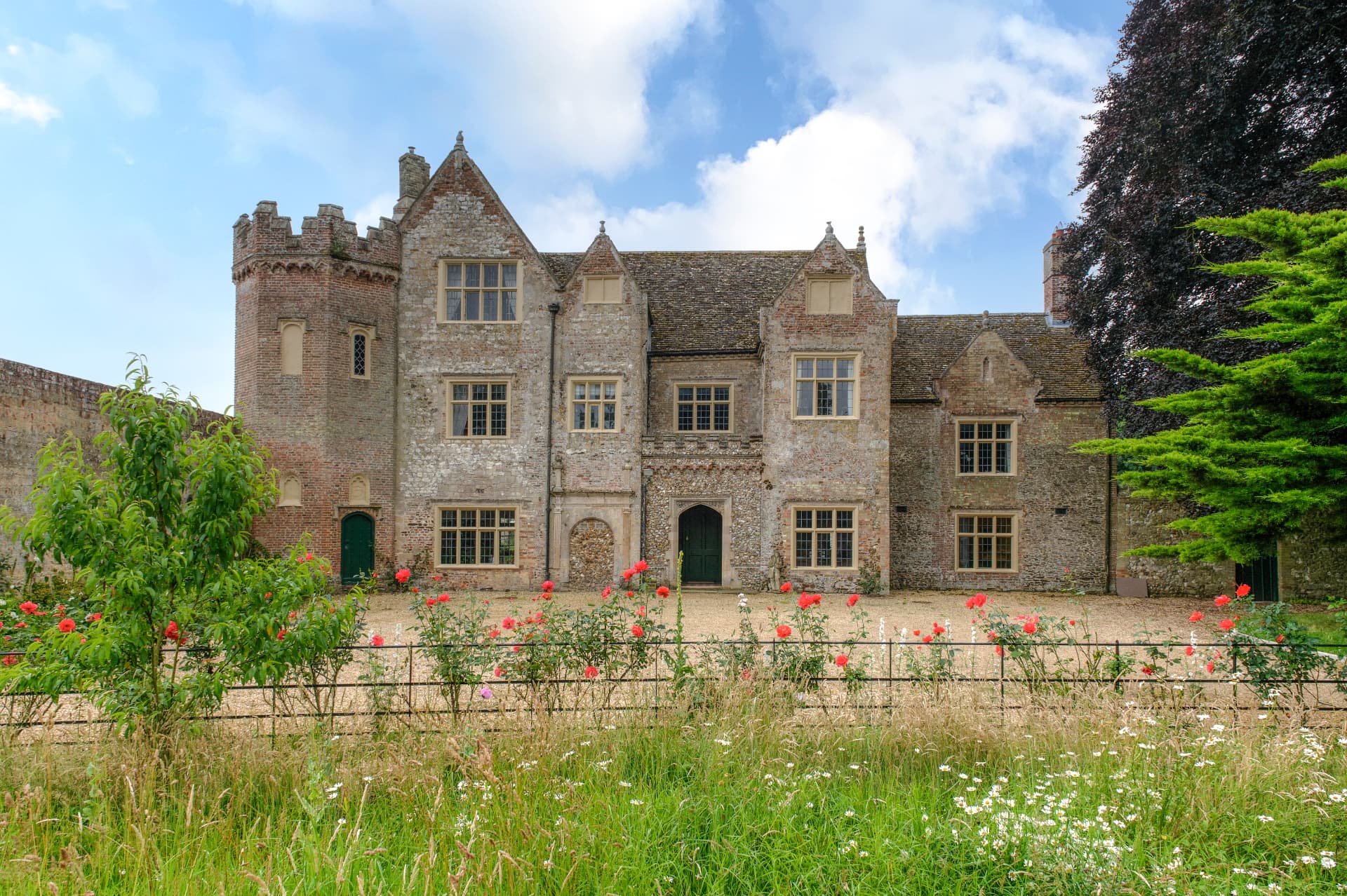 A flint and stone manor house with a castellated brick tower, mullioned windows and a red front door, seen beyond a rose-lined meadow