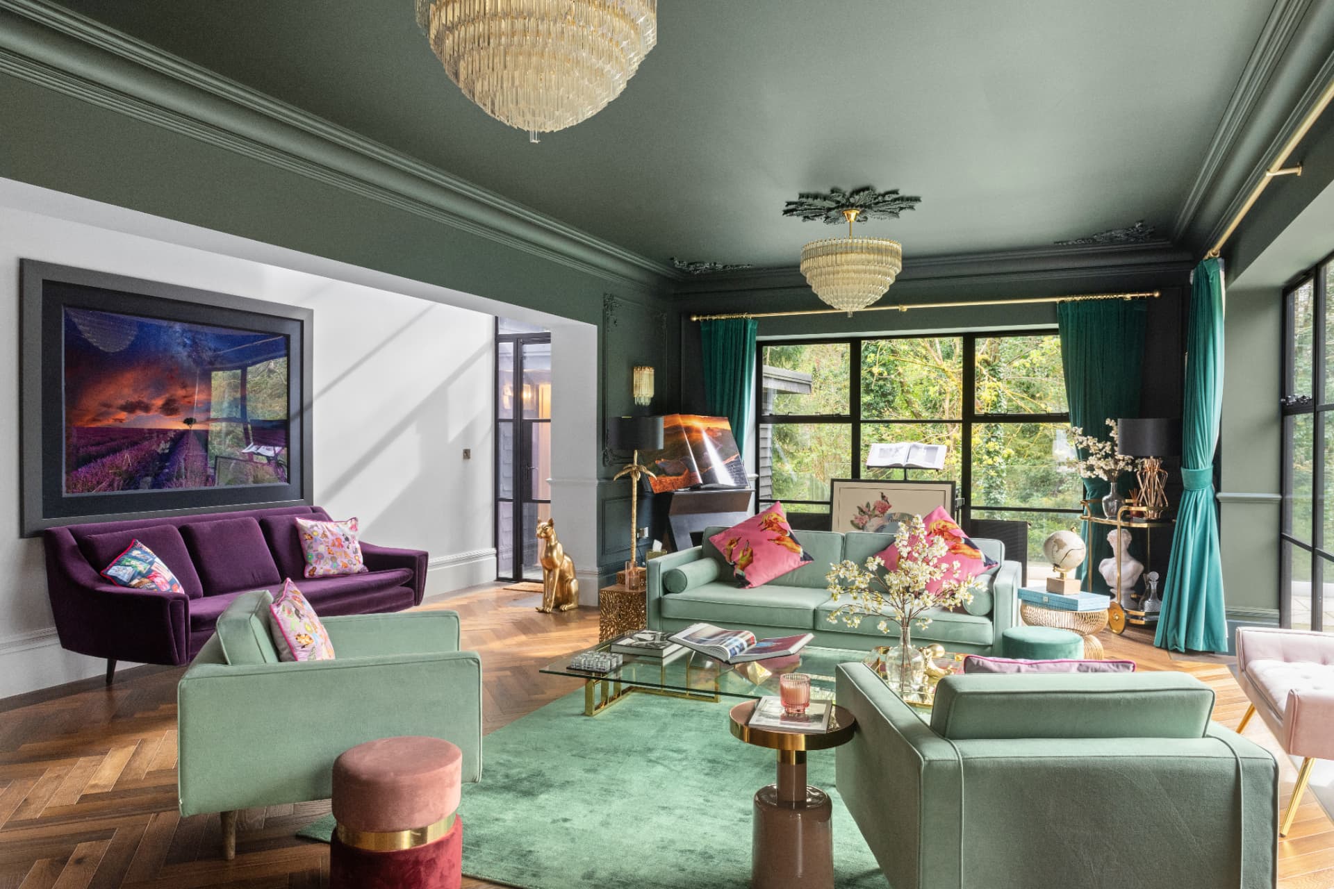 A drawing room in deep sage with a green-painted ceiling, two crystal chandeliers, velvet sofas in mint and plum, herringbone parquet and tall black-framed windows onto trees