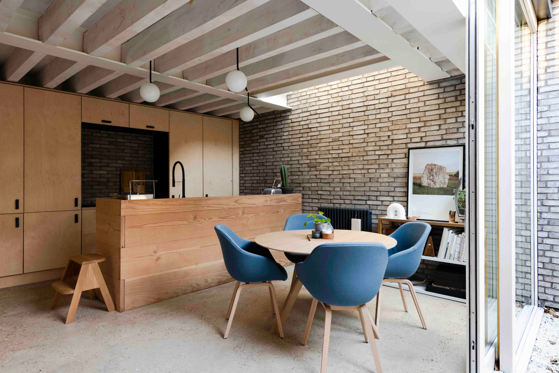 A pared-back kitchen and dining space with birch plywood joinery, an exposed brick wall and painted timber joists, blue tub chairs around a round oak table and glazing open to a courtyard