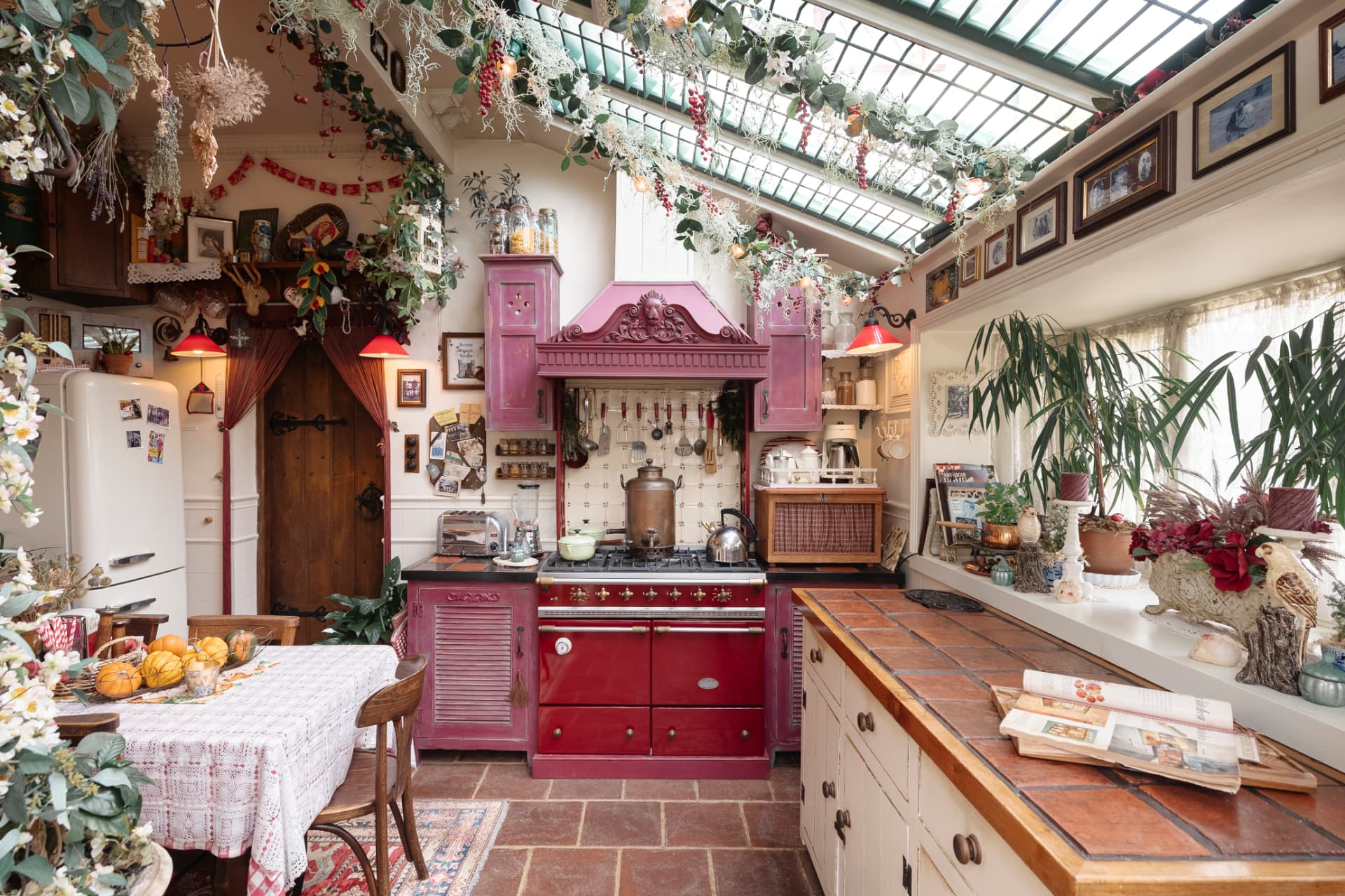 A country kitchen beneath a glazed roof, with a deep pink range surround, terracotta floor and dried flowers hung from the ceiling