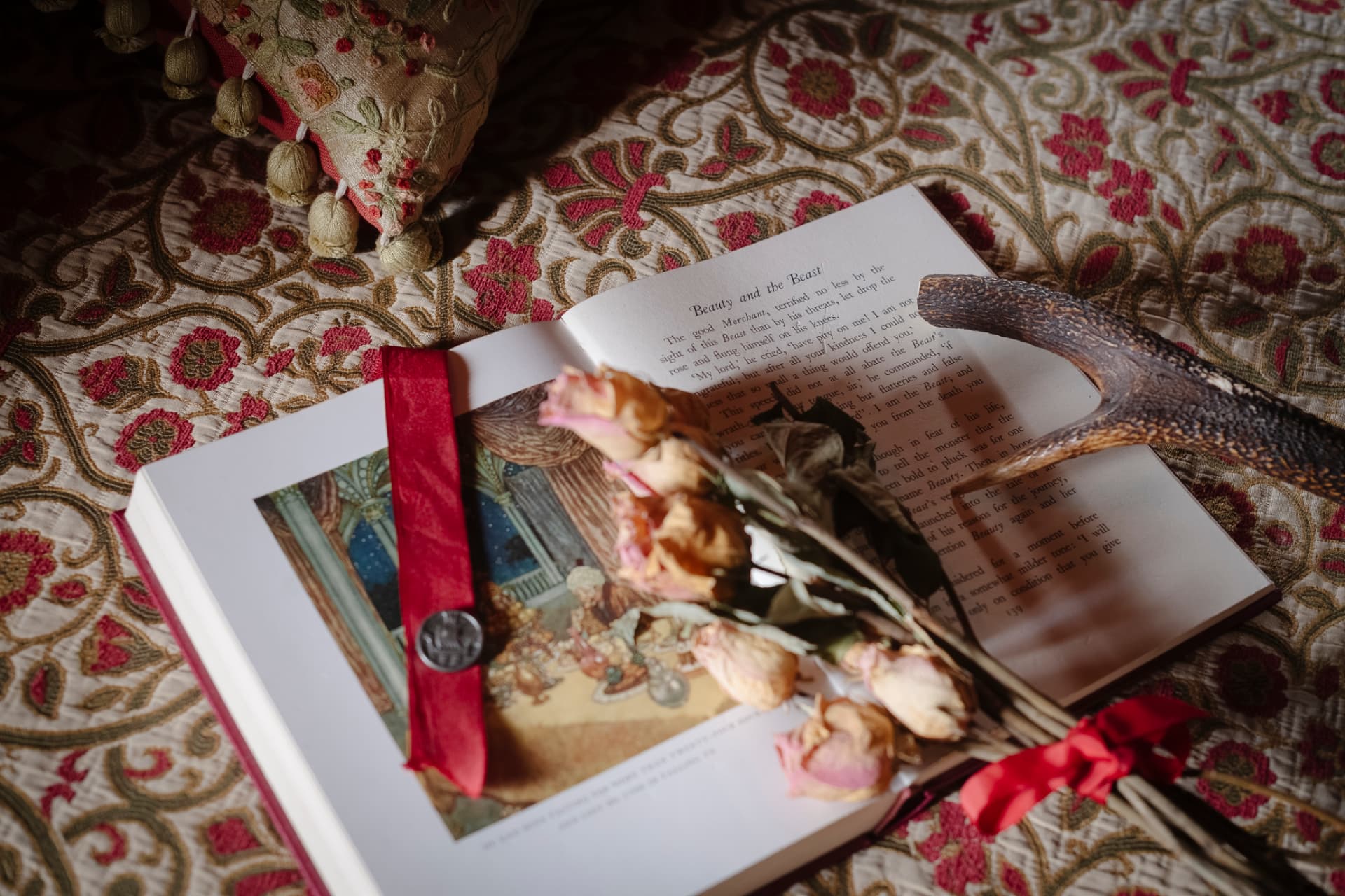 An open illustrated book resting on patterned fabric, a red ribbon marker and dried roses laid across the page