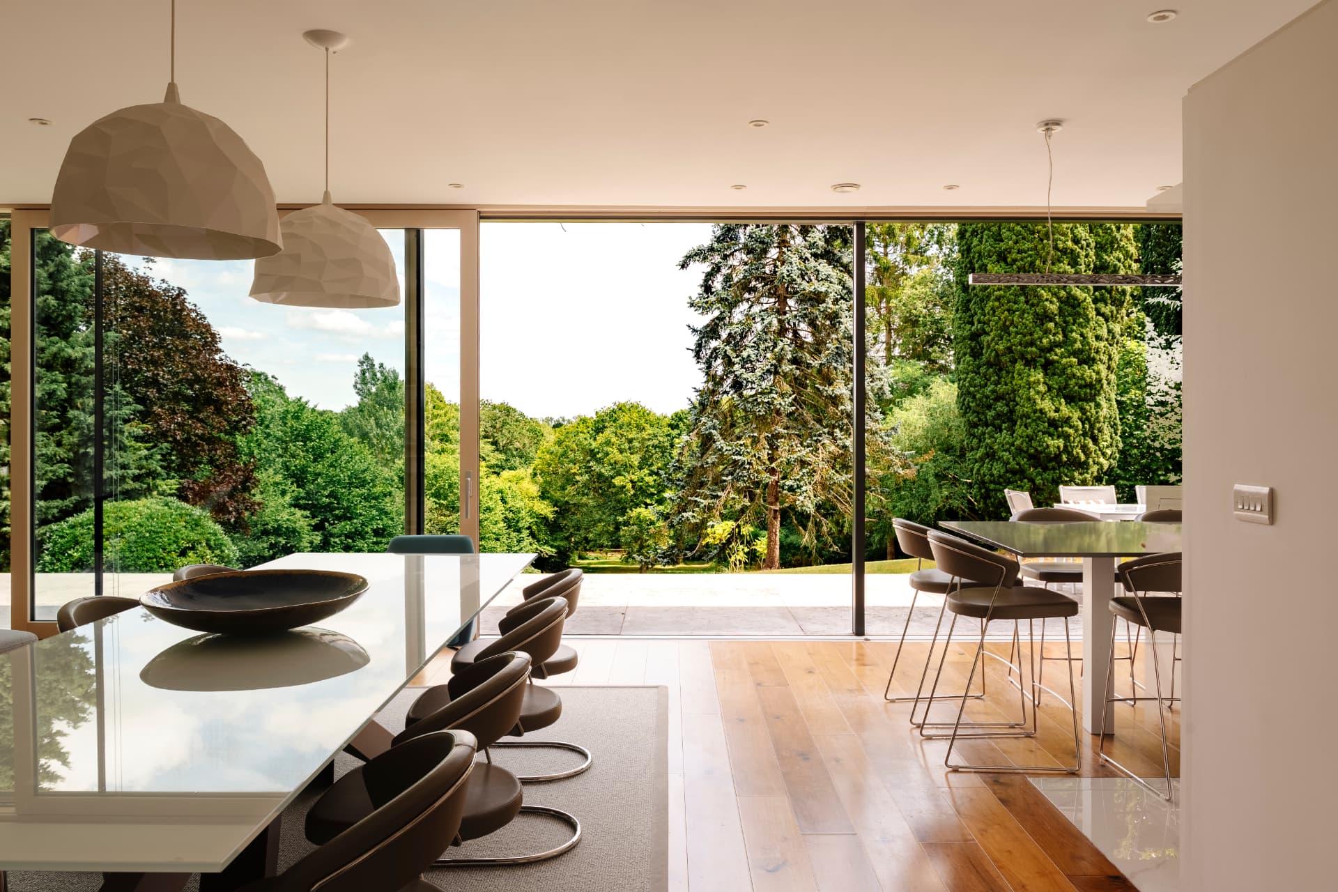 A dining room with sculptural pendants over a glass table, full-height sliding glazing pushed open onto a stone terrace and mature trees beyond