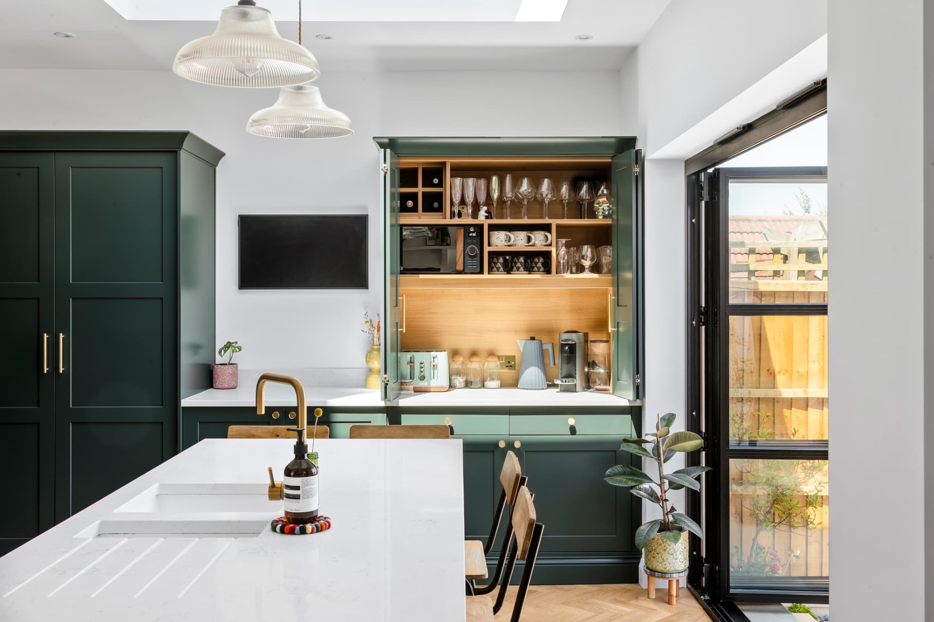 A deep green kitchen with a white quartz island, ribbed glass pendants, an open breakfast cupboard and glazed doors to a small garden