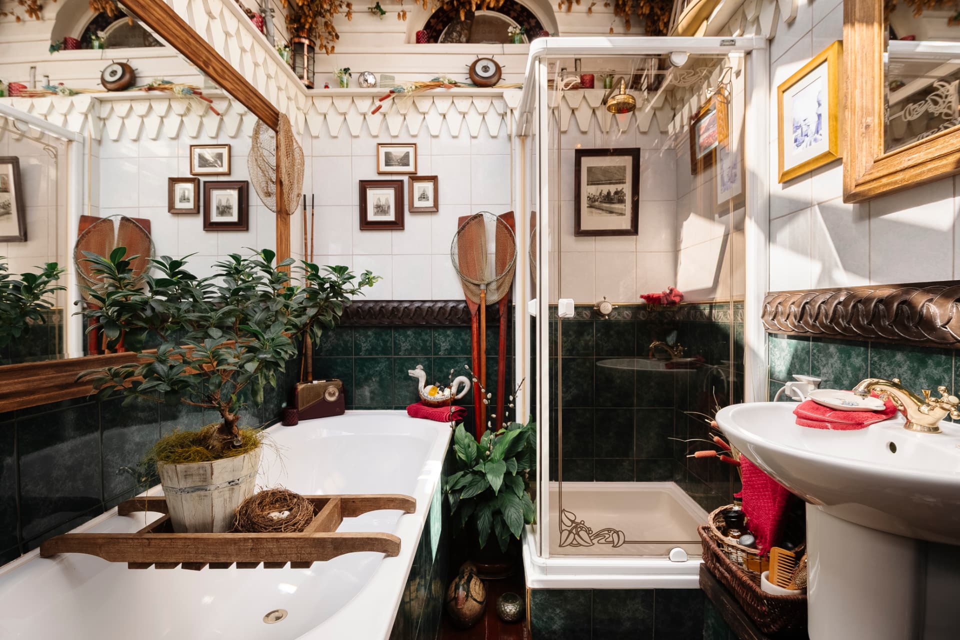 A characterful bathroom in white and dark green tile, with a bonsai and landing nets beside the bath, framed prints and brass fittings catching low sunlight