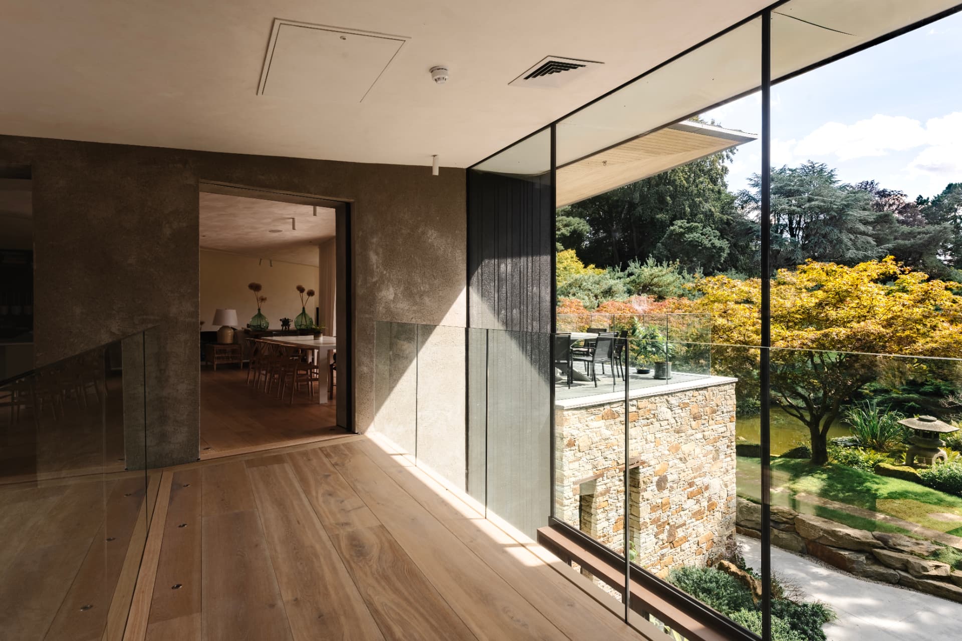 An oak-floored upper landing wrapped in full-height glazing, looking out over a stone-clad wing and a Japanese-style garden