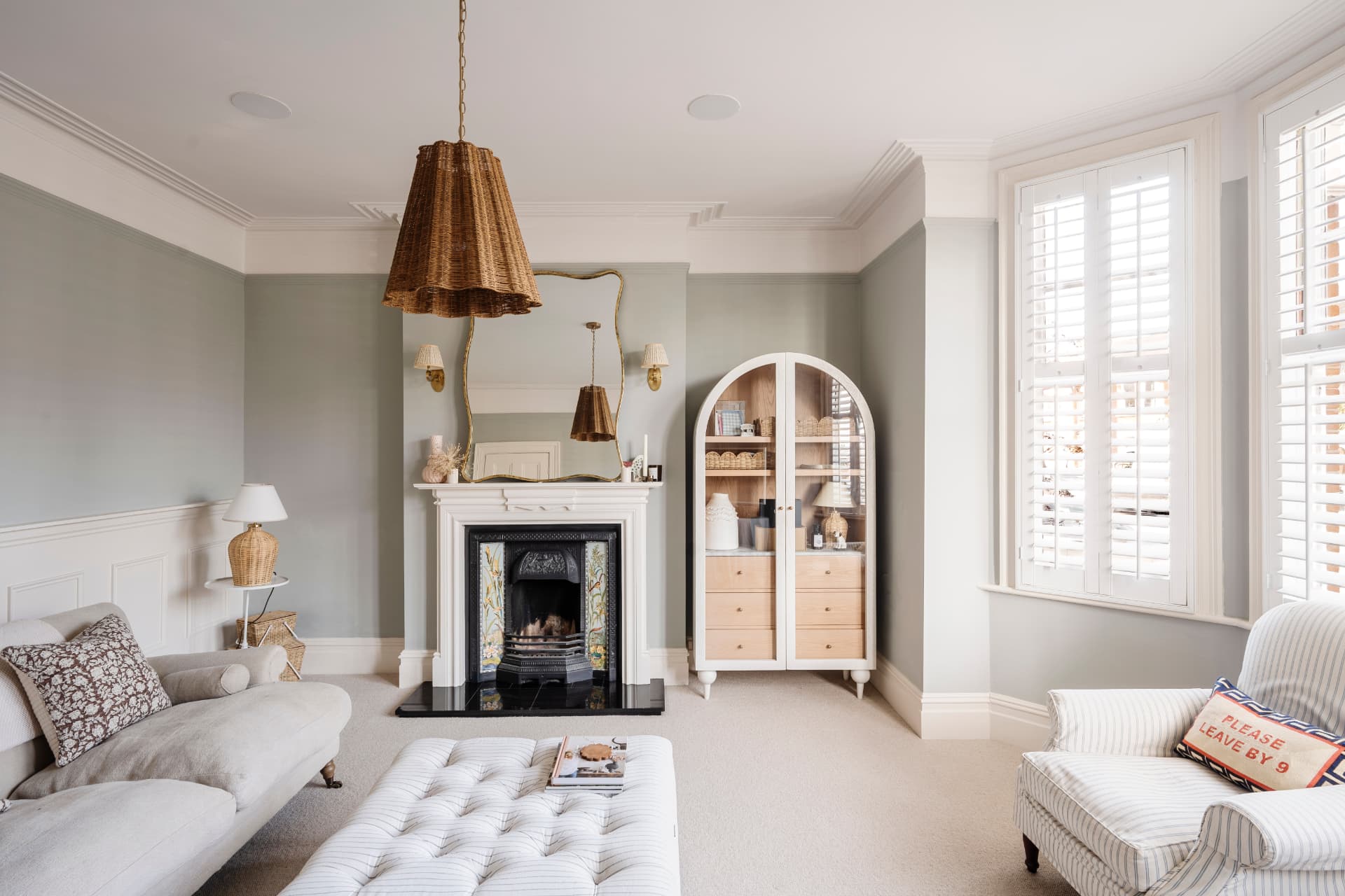 A sage-green sitting room with a tiled cast-iron fireplace, gilt mirror, wicker pendant and plantation shutters at a bay window