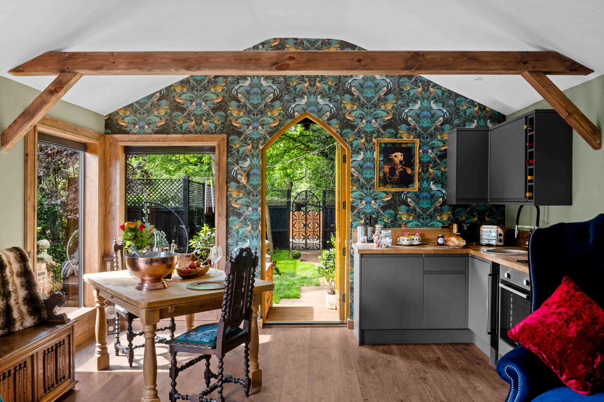 A timber-framed room with bold botanical wallpaper around an arched opening, dark kitchen units and a dining table, with a doorway open to the garden