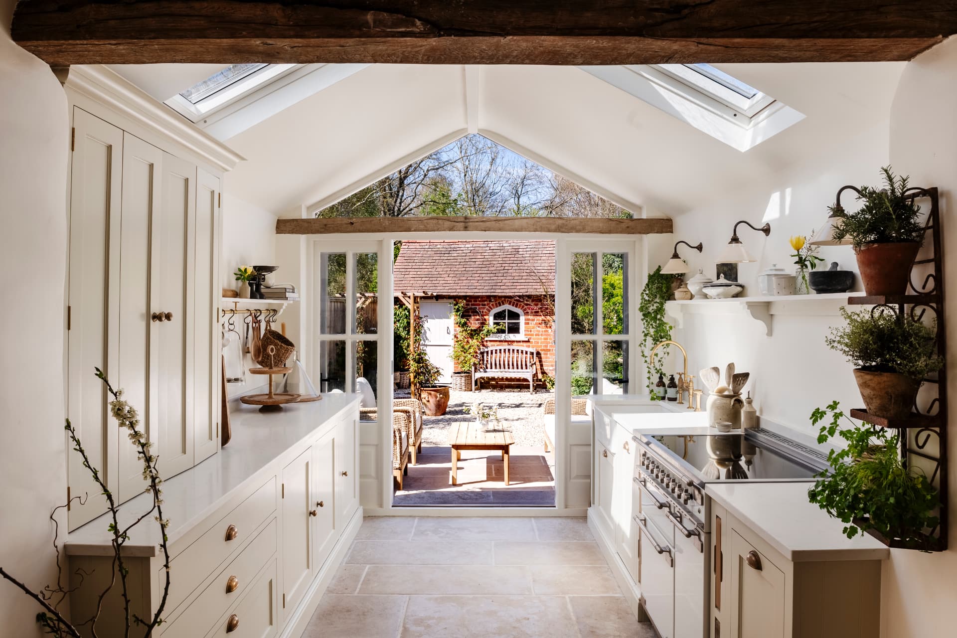 A narrow beamed kitchen under rooflights, cream cabinetry and potted herbs framing open garden doors onto a brick courtyard