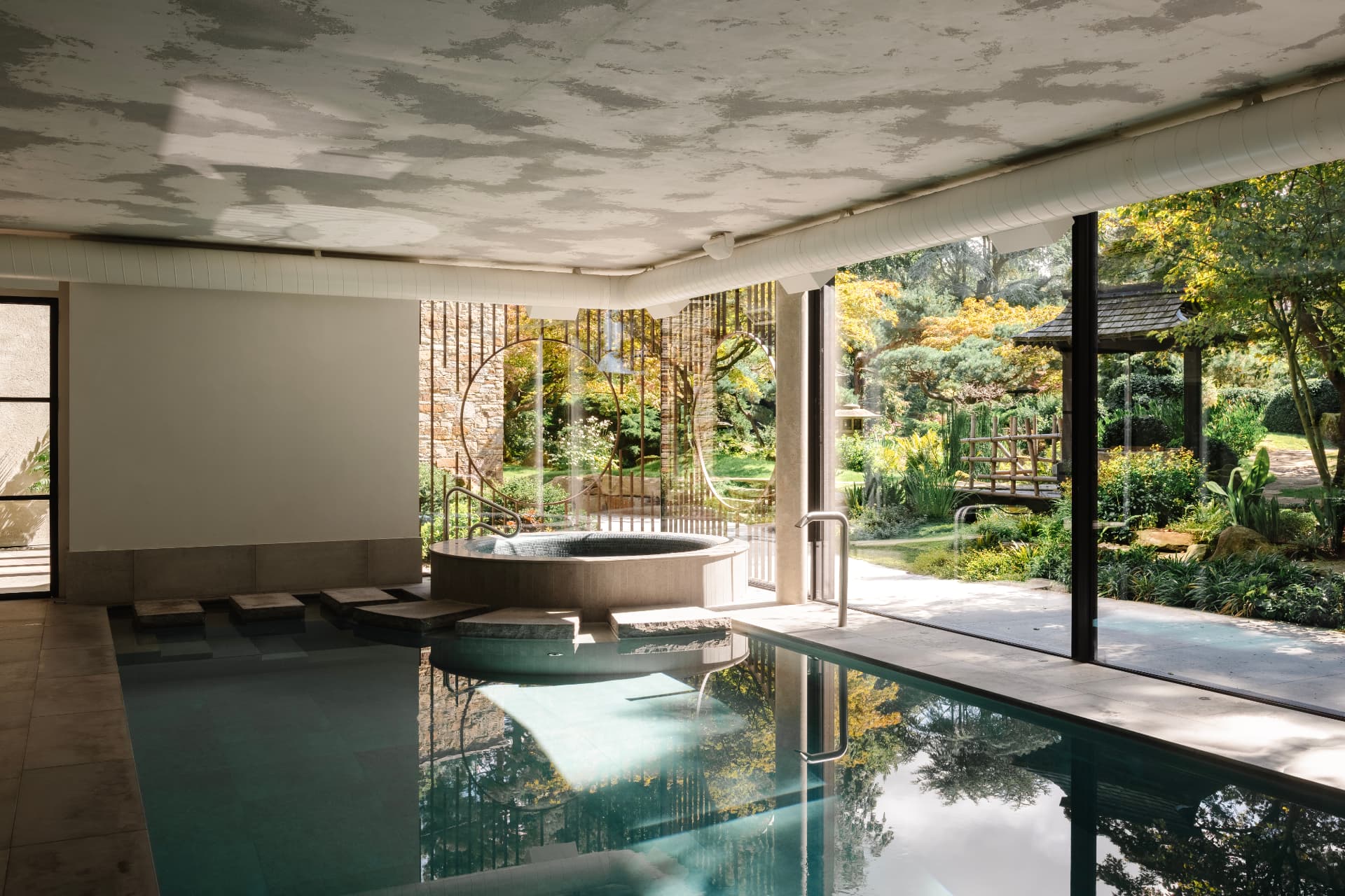 An indoor pool with still water reflecting a dappled ceiling, a circular stone planter at the water's edge and full-height glazing onto a wooded garden