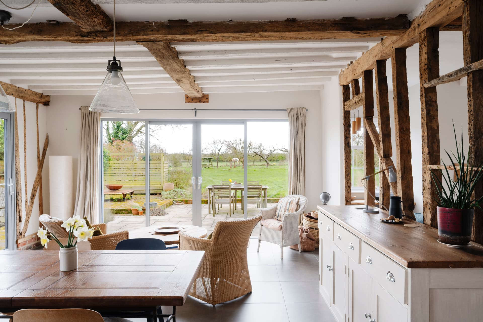 An oak-frame farmhouse interior with exposed timber posts and beams, a pendant light above a dining table, and sliding glazing opening onto open fields