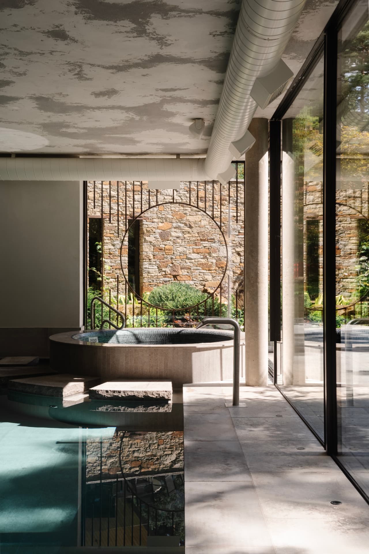 An indoor pool and circular spa beside full-height glazing, with a dry-stone wall and circular metal screen in the garden beyond