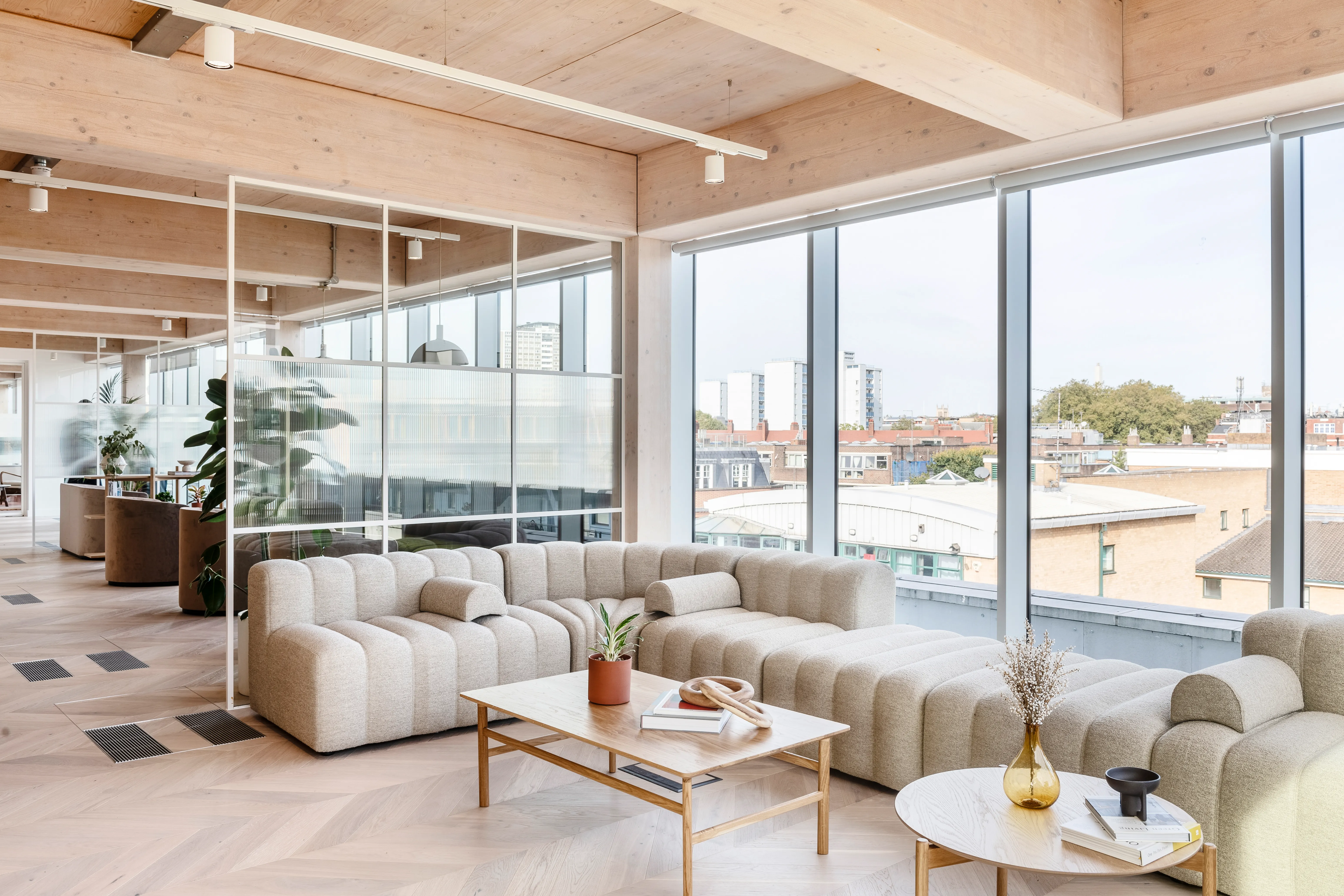 A bright office breakout area with floor-to-ceiling glazing over a city skyline, a plush modular sofa and a low coffee table