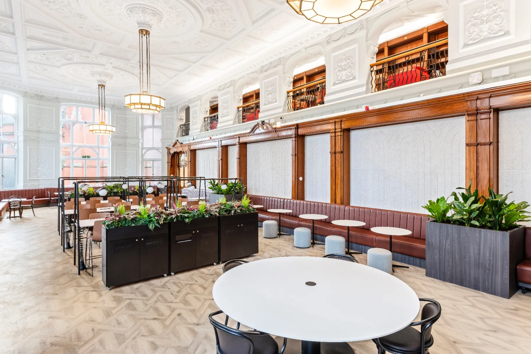 A grand double-height library hall with arched mullioned windows, ornate plasterwork cornicing and wood-panelled bays, furnished with reading tables and pendant lighting